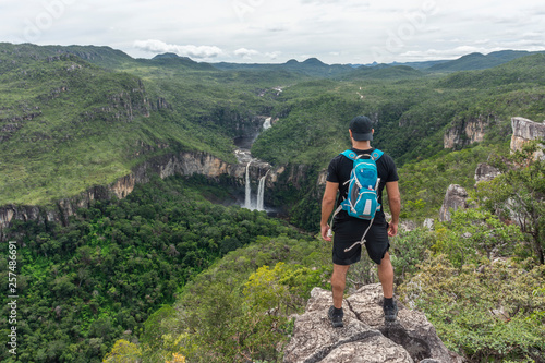 Young adult hiker on rocky edge looking at beautiful cerrado landscape with waterfalls and river in the green forest, Mirante da Janela peak, Chapada dos Veadeiros, Goias state, central Brazil