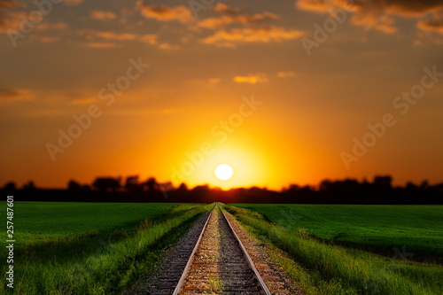 A railroad track dividing lush green agriculture fields under a bright orange and yellow sunset