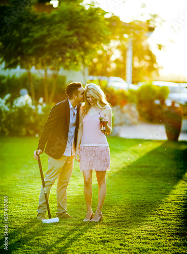 A young man stands with a croquet mallet in his one hand kisses his lady partner on her head. photo