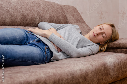 Blonde girl lying on the couch in blue jeans and a gray sweater, feels a pain in her stomach