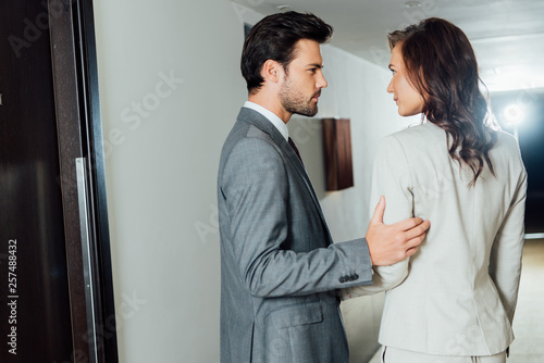 confident businessman touching hand of attractive businesswoman in formal wear while standing in hotel corridor