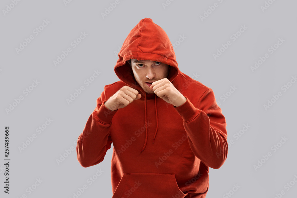 combat, aggression and people concept - young man in red hoodie fighting with fists or boxing over grey background