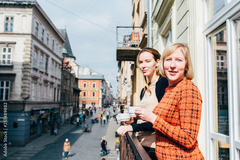 Portrait of two pretty blonds women, mother and daughter, drinking tea. Mature middle age and adult women standing on balcony and talking. Different generation parent concept