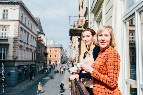 Portrait of two pretty blonds women, mother and daughter, drinking tea. Mature middle age and adult women standing on balcony and talking. Different generation parent concept
