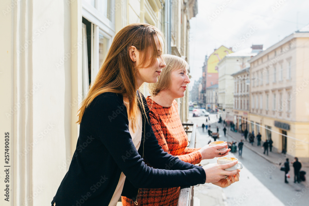 Fototapeta premium Portrait of two pretty blonds women, mother and daughter, drinking tea. Mature middle age and adult women standing on balcony and talking. Different generation parent concept