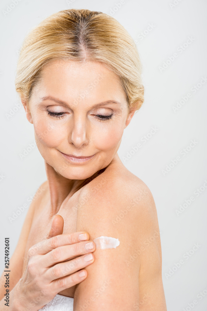 beautiful and mature woman looking down and applying cosmetic cream on shoulder isolated on grey beautiful and mature woman looking down and applying cosmetic cream on shoulder isolated on grey