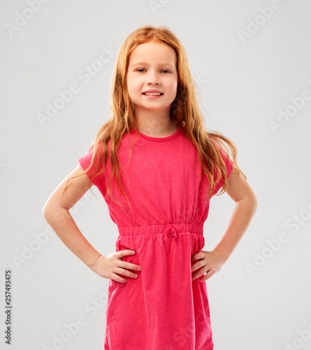 childhood and people concept - smiling red haired girl posing in pink dress over grey background
