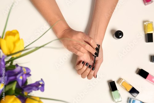 Woman applying nail polish near bottles on white table with flowers, top view