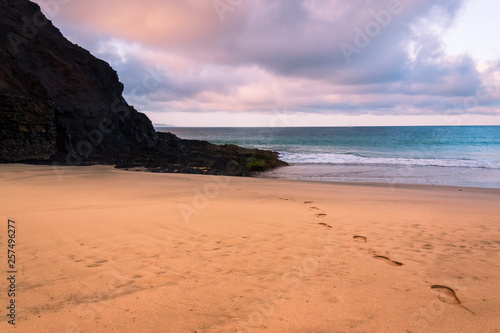 Foot prints in the sand on an empty beach leading towards the turquoise sea water under a beautiful sunrise sky. The Wax Beach (Playa de la Cera) tourist attraction in Lanzarote, Canary Islands. 