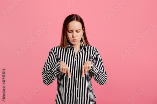 Sad upset young woman point down. She stand in studio and look. Isolated over pink background.