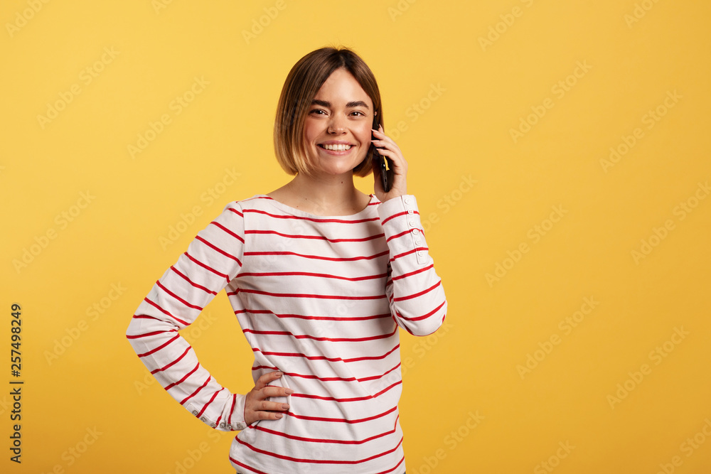 Nice cheerful young woman smile and pose on camera. She talk on phone. Isolated over yellow background. Nice cheerful young woman smile and pose on camera. She talk on phone. Isolated over yellow background.