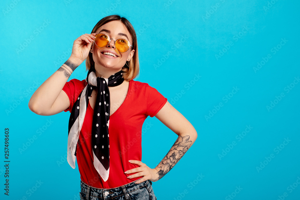 Cheerful positive nice young woman pose on camera and smile. She hold hand on yellow sunglasses. Shawl on head. Isolated over blue background.