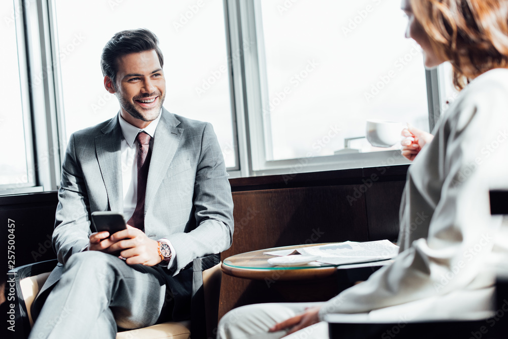 selective focus of cheerful businessman holding smartphone and looking at businesswoman with cup