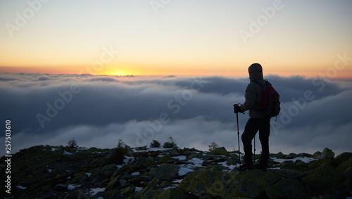 Back view of tourist hiker in warm jacket with backpack and trekking sticks on mountain peak on background of valley covered with white clouds stretching to horizon and bright morning sky at sunrise.