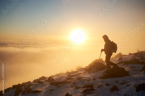 Silhouette of tourist hiker with backpack and trekking sticks hiking on rocky snowy mountain steep slope on background of foggy valley filled with white puffy clouds, raising sun and blue sky at dawn.