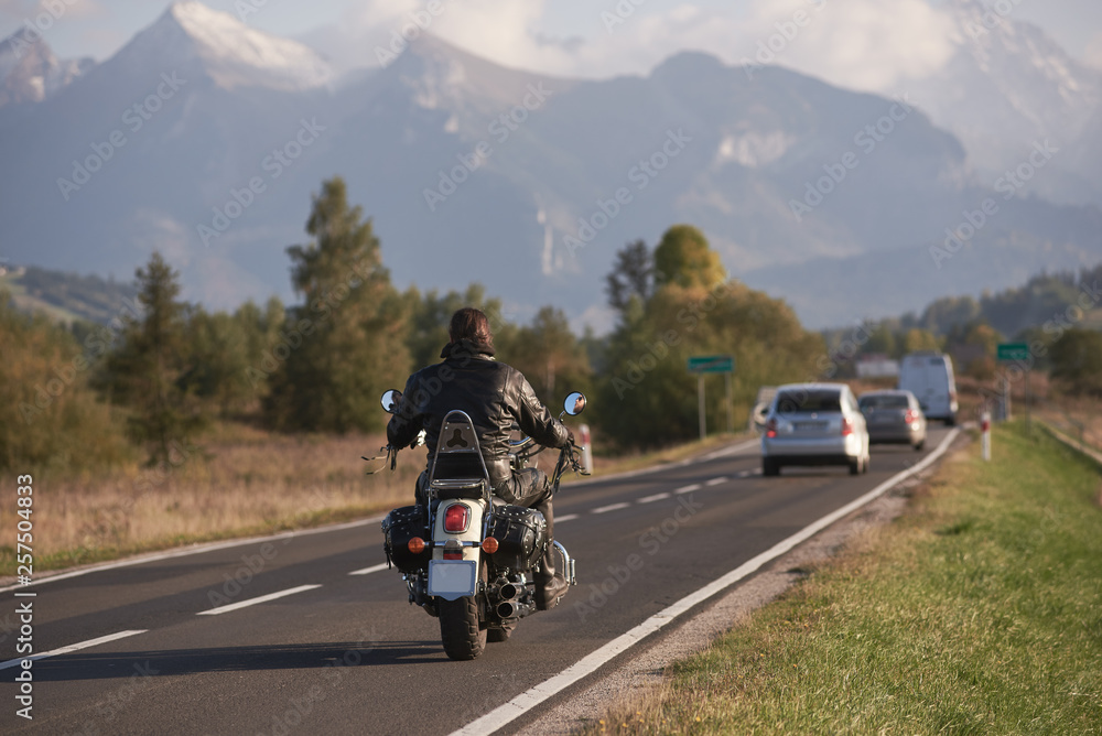 Back view of motorcyclist in black leather jacket riding motorcycle along road on blurred background of beautiful mountain range with snowy peaks, moving vehicles on bright sunny day.