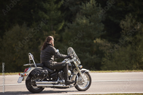 Side view of bearded long-haired motorcyclist in sunglasses and black leather clothing riding motorbike along narrow asphalt path on sunny summer day on background of tall trees.