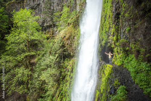 Columbia River Gorge, OR, USA. A man stands at the opening of a tunnel along a lush green, moss-lined trail with a waterfall falling from above. photo