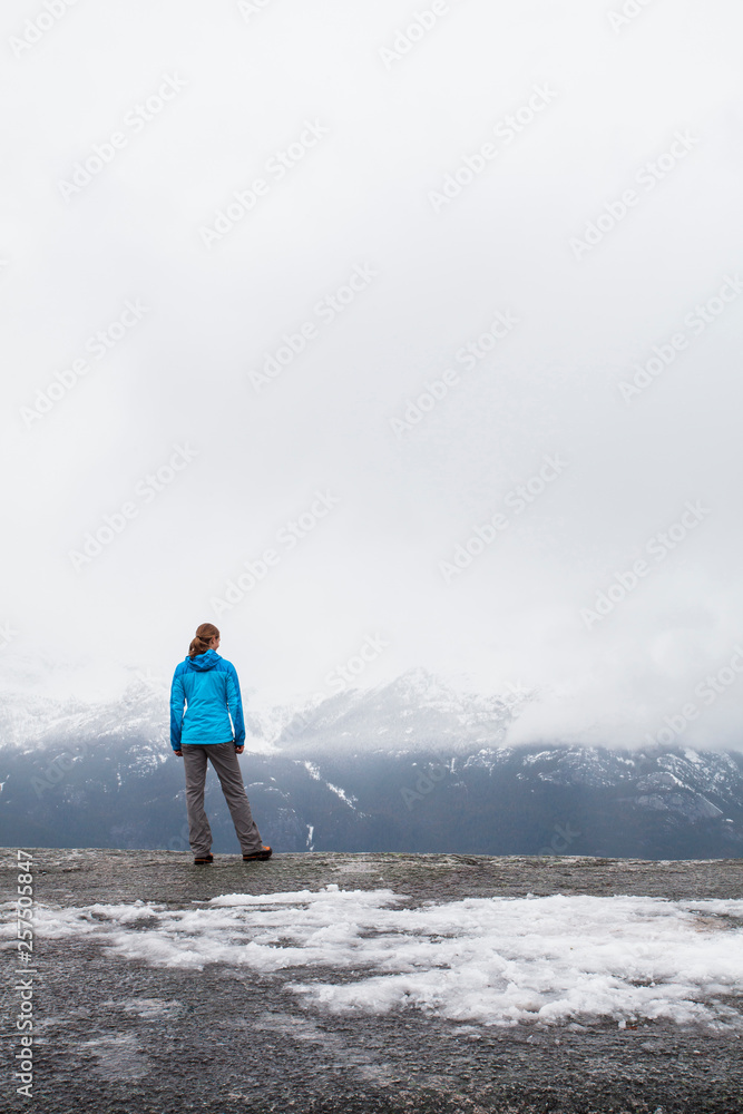 SQUAMISH, BRITISH COLUMBIA, CANADA. A young woman in hiking clothes looks over a foggy, wintry view from a flat rock edge. SQUAMISH, BRITISH COLUMBIA, CANADA. A young woman in hiking clothes looks over a foggy, wintry view from a flat rock edge.