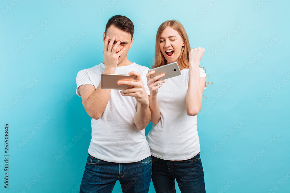 Young excited couple standing , playing games on mobile phones, on a blue background