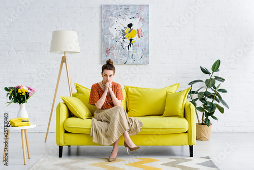 Quadro su tela pensive elegant young woman sitting with crossed legs and clenched hands on yell