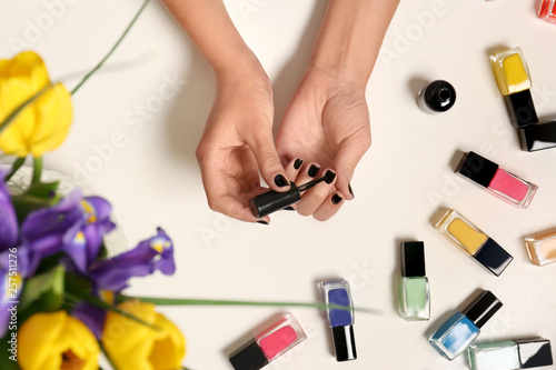Woman applying nail polish near bottles on white table with flowers  top view