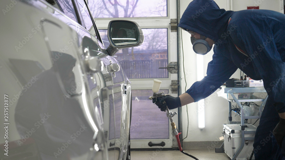 In slow motion, a Man (Man) professional in working uniform with spray guns in his hands and a respirator in protective glasses puts paint (ceramics) on the car on different parts of the car. In slow motion, a Man (Man) professional in working uniform with spray guns in his hands and a respirator in protective glasses puts paint (ceramics) on the car on different parts of the car.