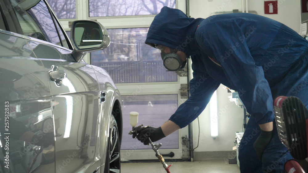 In slow motion, a Man (Man) professional in working uniform with spray guns in his hands and a respirator in protective glasses puts paint (ceramics) on the car on different parts of the car. In slow motion, a Man (Man) professional in working uniform with spray guns in his hands and a respirator in protective glasses puts paint (ceramics) on the car on different parts of the car.