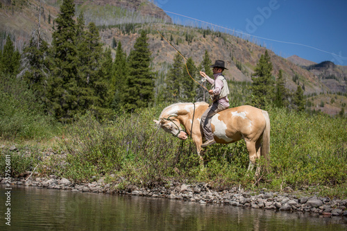 A man fishes from the back of a pinto horse in cowboy hat in Montana backcountry near Yellowstone.