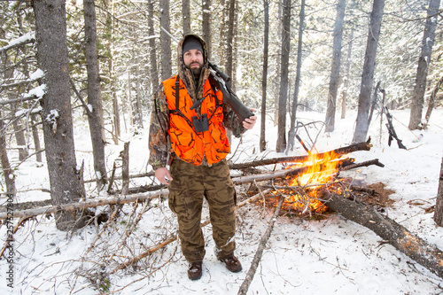A male hunter in camouflage with a blaze orange vest on holds his rifle next to a fire he started when the weather turned colder during an elk and deer hunting trip in Northwest Montana.