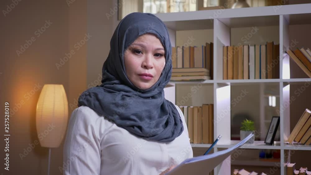 Closeup shoot of adult muslim businesswoman studying the graph in the library being thoughtful and looking at camera on the workplace indoors Closeup shoot of adult muslim businesswoman studying the graph in the library being thoughtful and looking at camera on the workplace indoors