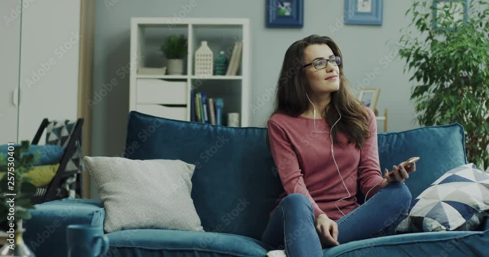 Caucasian attractive young lady sitting on the sofa and resting while enjoying music on the smartphone via headphones at home. Caucasian attractive young lady sitting on the sofa and resting while enjoying music on the smartphone via headphones at home.