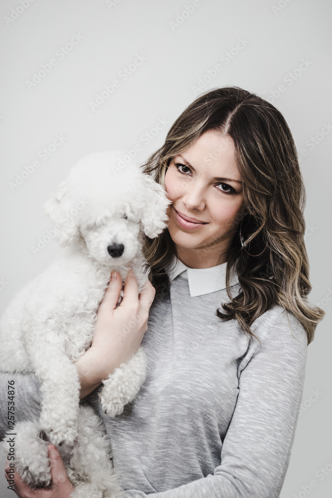 Beautiful young woman smiling and holding gorgeous dwarf white poodle dog. She standing against white wall and looking at camera. Beautiful young woman smiling and holding gorgeous dwarf white poodle dog. She standing against white wall and looking at camera.