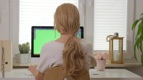 back view of woman sitting at white desk and working on computer keyboard in front of display with isolated green screen