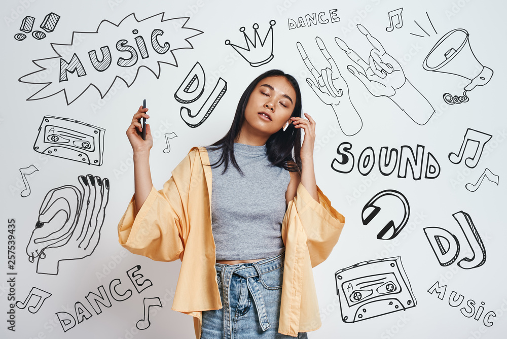 Enjoying her favourite song. Attractive and young asian woman in casual wear listening music with closed eyes while standing against grey background with music theme doodles. Enjoying her favourite song. Attractive and young asian woman in casual wear listening music with closed eyes while standing against grey background with music theme doodles.
