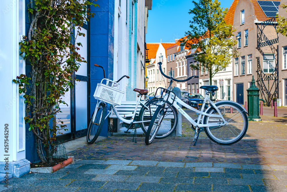 Two white romantic bicycles standing near each other and parked in the shadow of the street. Outdoor activities. Summer concept. Vacation. Travel. Two white romantic bicycles standing near each other and parked in the shadow of the street. Outdoor activities. Summer concept. Vacation. Travel.