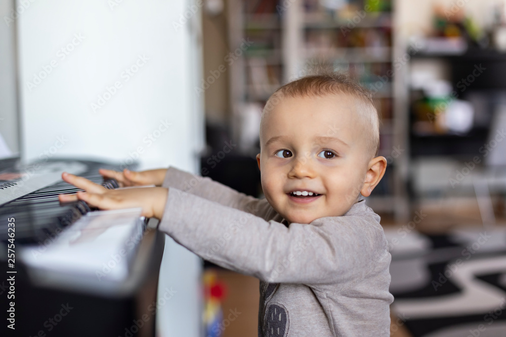 A toddler boy is standing at the piano and smiling. Baby boy wants to play piano at home, child likes classic music. A toddler boy is standing at the piano and smiling. Baby boy wants to play piano at home, child likes classic music.