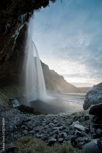 Seljalandsfoss waterfall in Iceland sprays mist onto rocks, which has frozen into crystals, as seen from behind the waterfall.