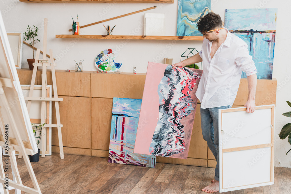 barefoot artist in white shirt and blue jeans carrying paintings in gallery