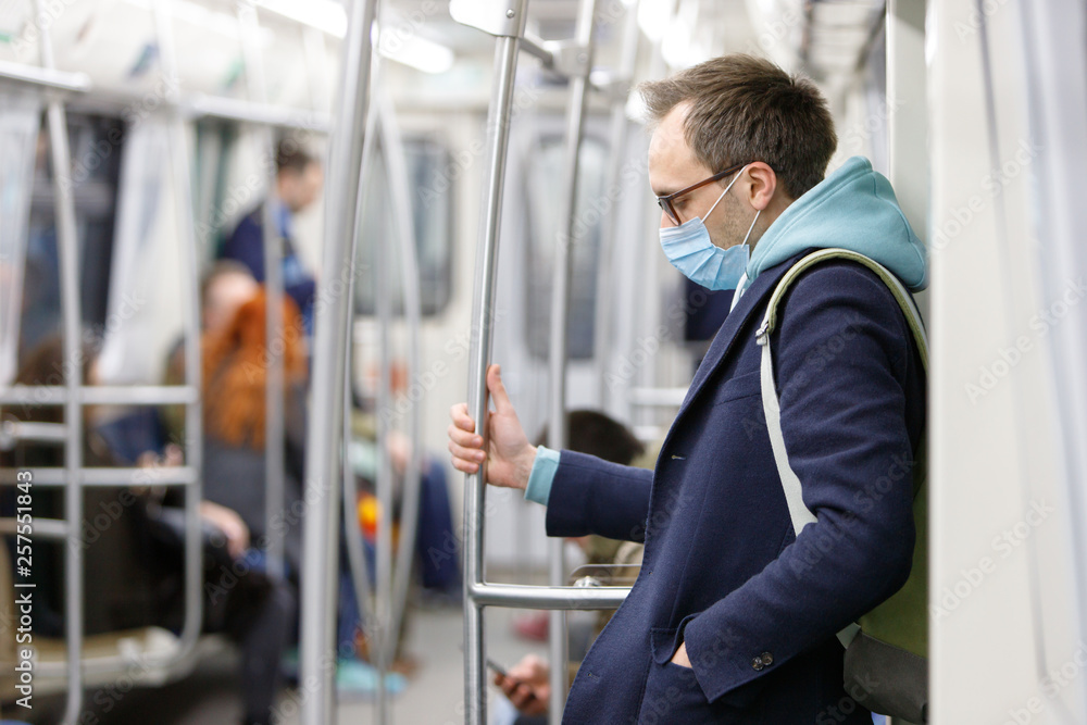 Exhausted man in eyeglasses feeling sick, wearing protective mask against transmissible infectious diseases and as protection against the flu in public transport/subway, people on background. 