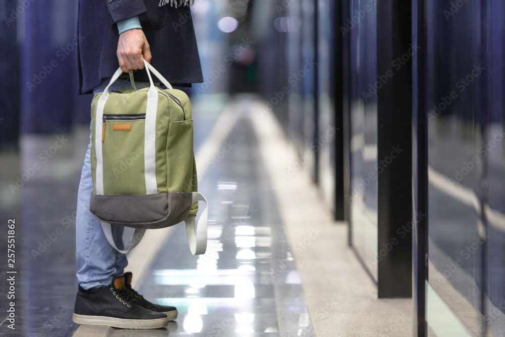 Obraz premium Closeup of man waiting train at metro station, holding a stylish light green cloth backpack, soft focus, empty subway platform on background, side view. Hand luggage concept