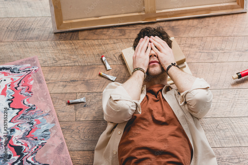 Obraz premium overhead view of exhausted artist lying on wooden floor and holding hands on face