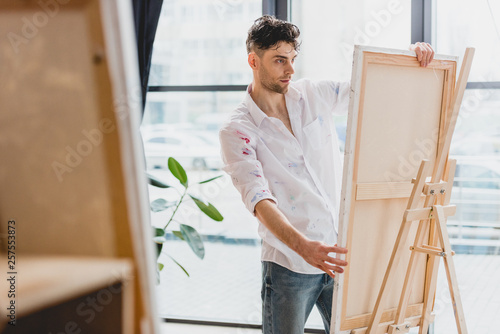 selective focus of handsome artist fixing canvas on easel in gallery