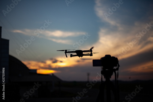 Silhueta de câmera fotográfica no tripé e drone capturando o por do sol, céu azul e laranja, com nuvens.