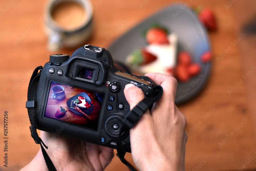 Hands of a photographer holding a camera with strawberry cake photo on the screen with the cake on the plate and a cup of coffee in the blurred background. Closeup. - obrazy, fototapety, plakaty 