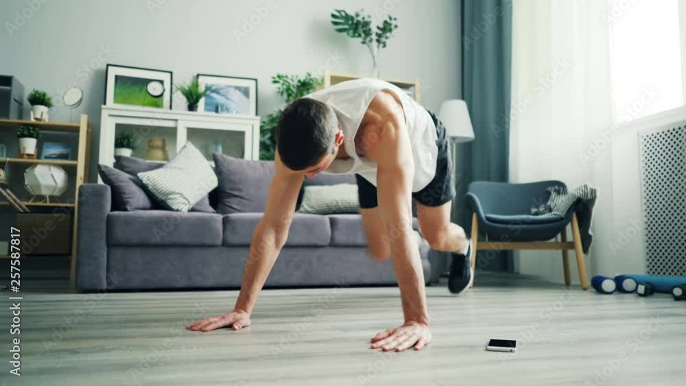 Strong young man in sportswear is doing cardiovascular exercises in plank position practising alone using smartphone. Domestic sports and modern technology concept. Strong young man in sportswear is doing cardiovascular exercises in plank position practising alone using smartphone. Domestic sports and modern technology concept.