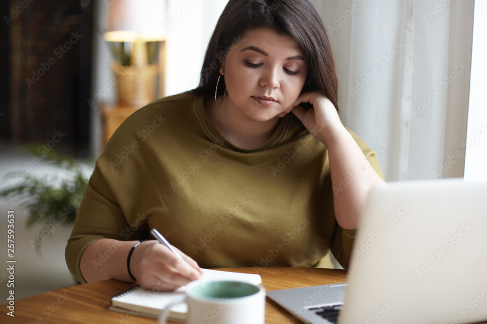 Attractive concentrated plus size young self employed woman with curvy body working remotely from home, making notes in copybook, planning business appointments. Communication, technology and gadgets Attractive concentrated plus size young self employed woman with curvy body working remotely from home, making notes in copybook, planning business appointments. Communication, technology and gadgets
