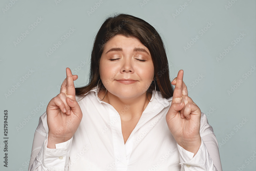 Emotional cute young plus size female with neat make up closing eyes and keeping fingers crossed, eager to get position in large successful company. Attractive chubby woman wishing for good luck Emotional cute young plus size female with neat make up closing eyes and keeping fingers crossed, eager to get position in large successful company. Attractive chubby woman wishing for good luck