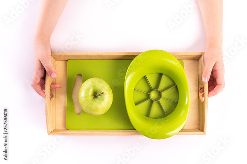 Children's hands hold a wooden tray with montessori materials for a lesson from the practical life zone. Apple slicer, cutting board,green apple. Isolate on white background.