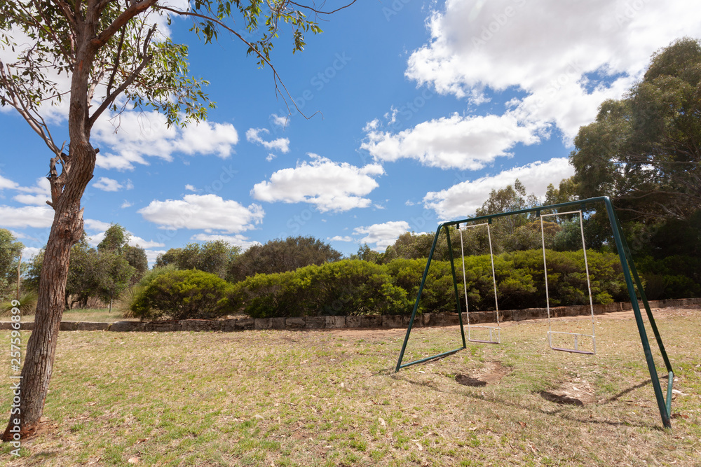 Two empty swings on grass with a small tree in the foreground under a blue sky with some white clouds Two empty swings on grass with a small tree in the foreground under a blue sky with some white clouds